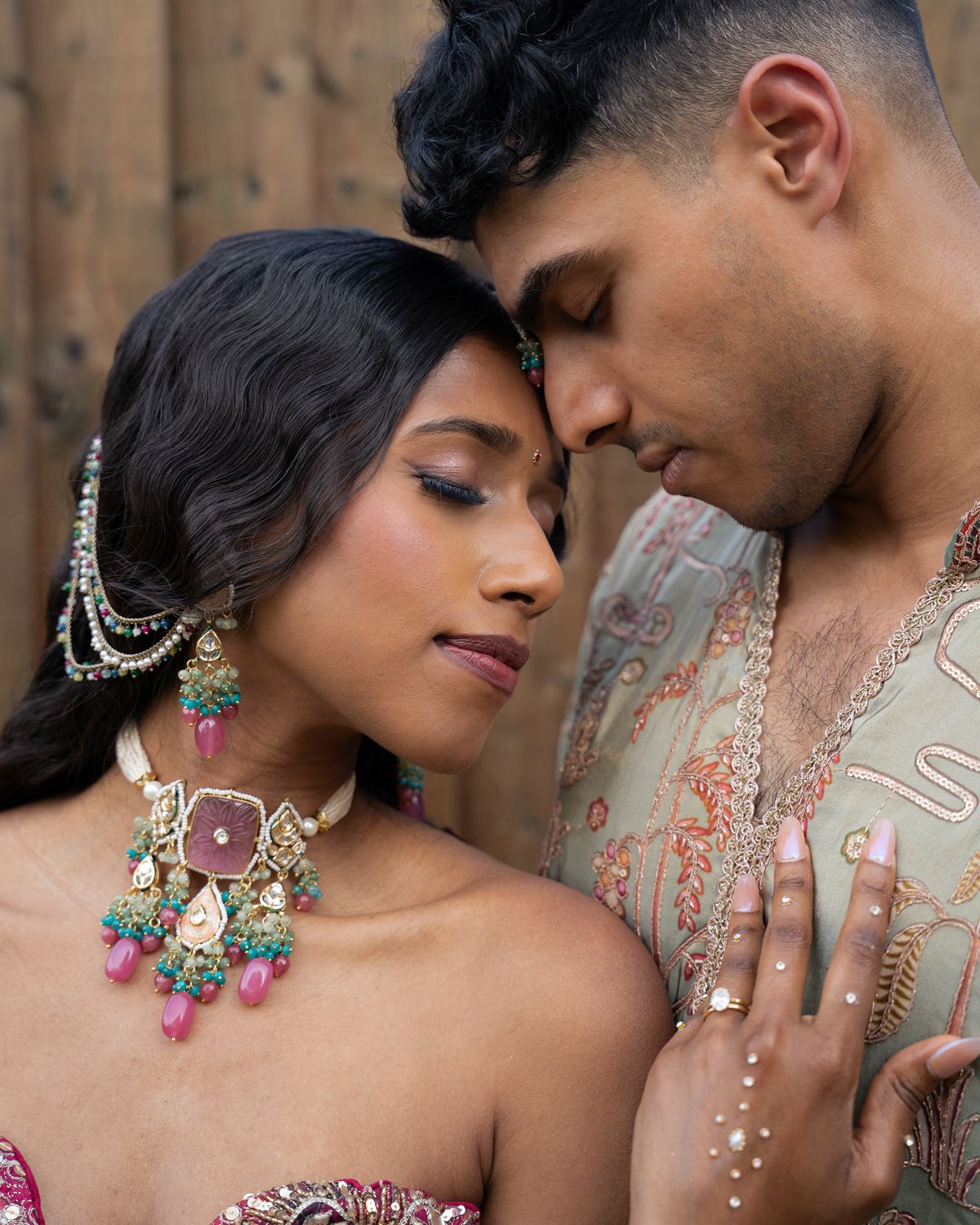 A Hindu bride and groom with foreheads touching, her pink and green kundan jewellery and his embroidered sage kurta close to camera
