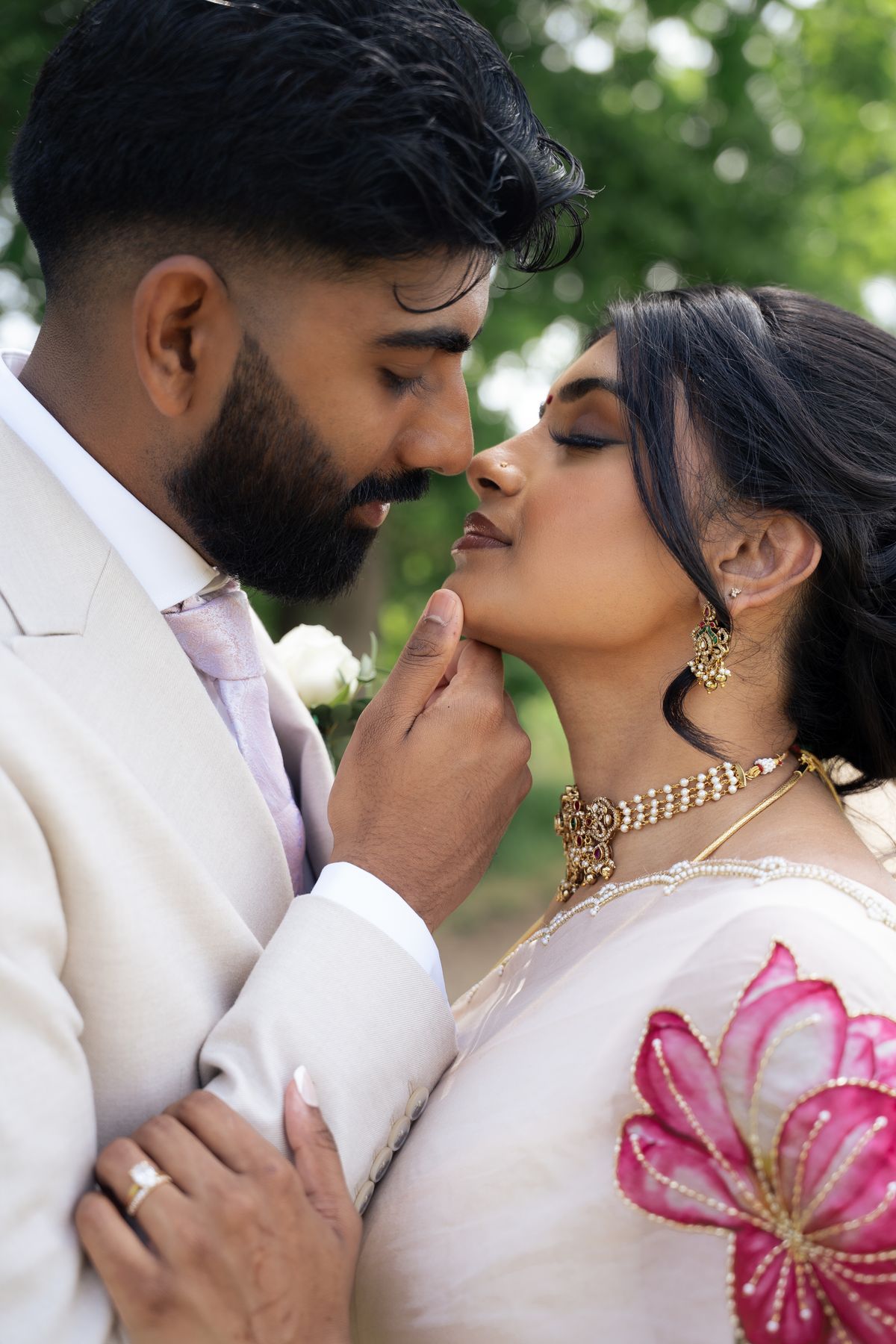 Civil wedding portrait: a groom in an ivory suit gently lifts his bride's chin, her pink lotus-embroidered saree and gold jewellery in soft light
