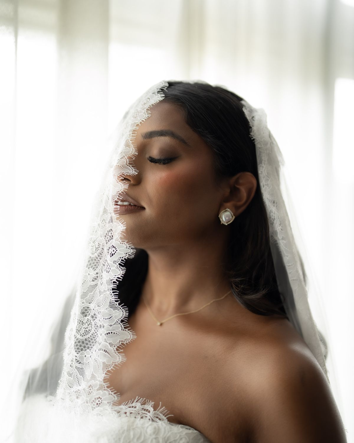 A bride with her eyes closed beneath a lace-edged veil, softly backlit by a window