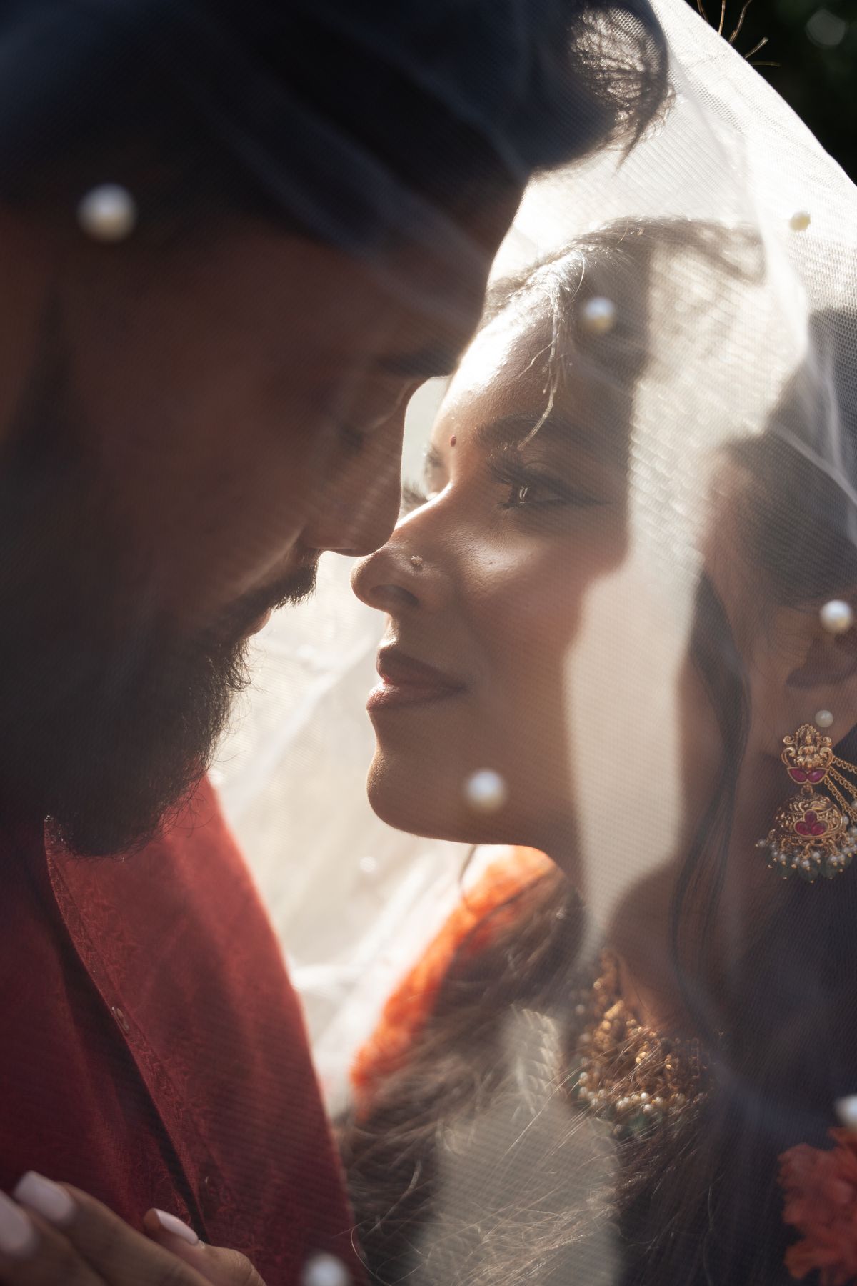 A couple nose to nose beneath a sheer pearl-dotted veil, backlit, her gold temple jhumkas catching the light