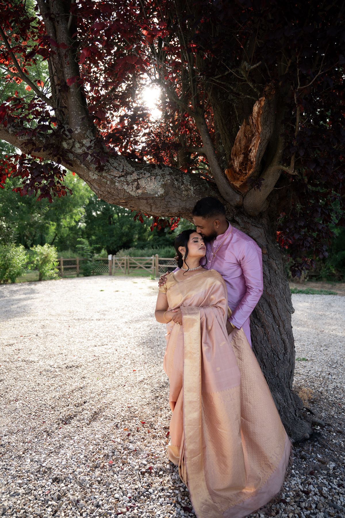 A couple leaning against a tree with deep red leaves, the bride in a blush silk saree and the groom in pink, with sunlight breaking through the branches