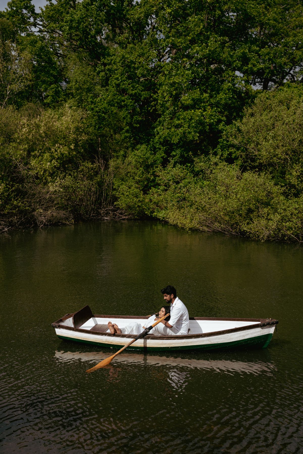 A couple resting together in a white rowing boat on a still, dark green lake, framed by trees