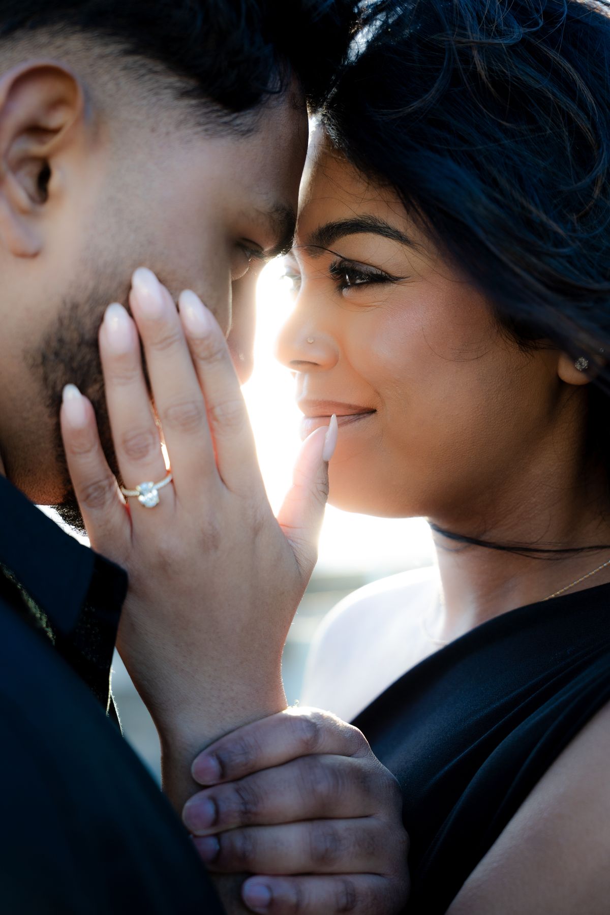 A couple with foreheads touching against bright backlight, her hand on his cheek showing her engagement ring