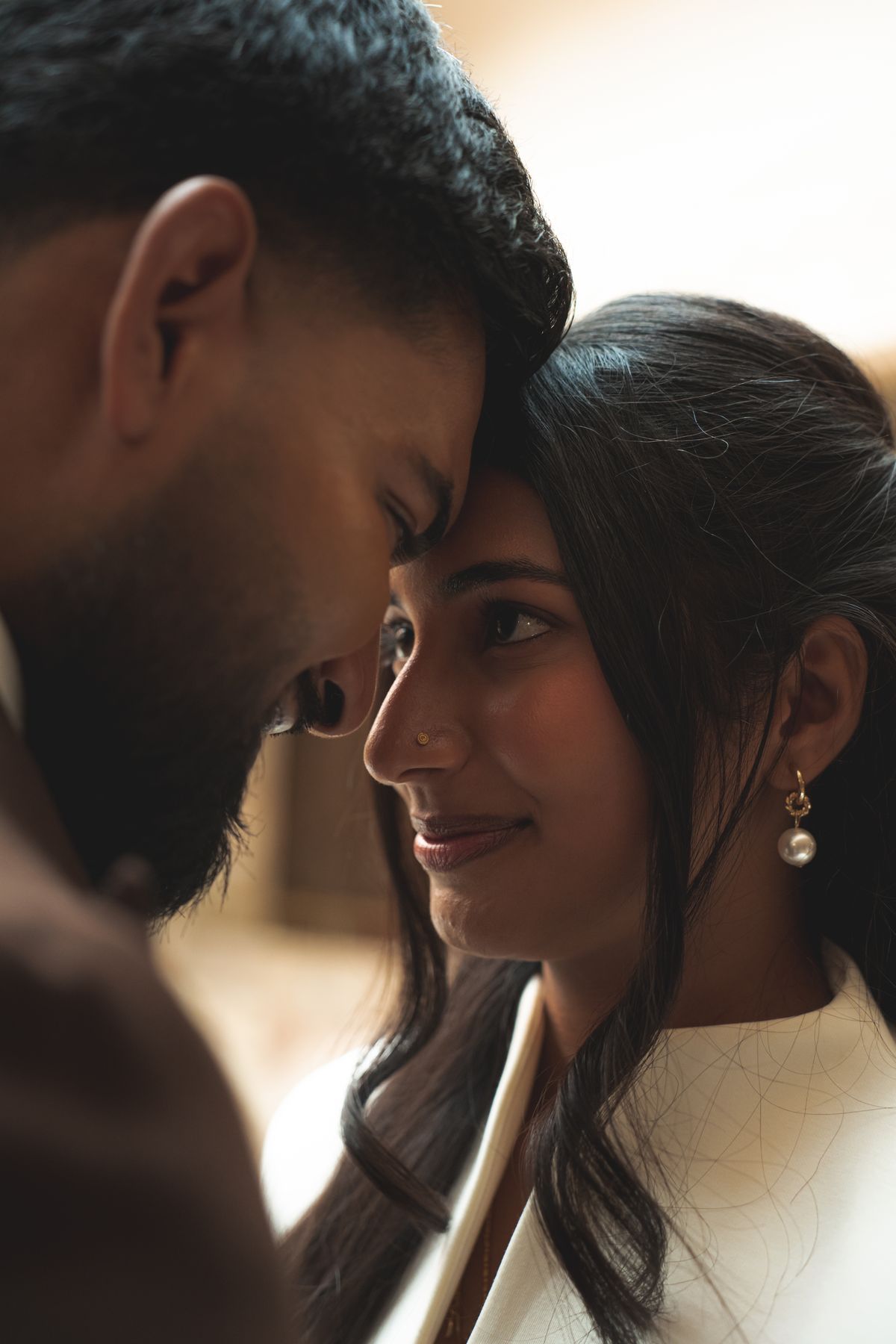 A couple with foreheads touching in warm light, the bride smiling softly in a white jacket and pearl drop earring