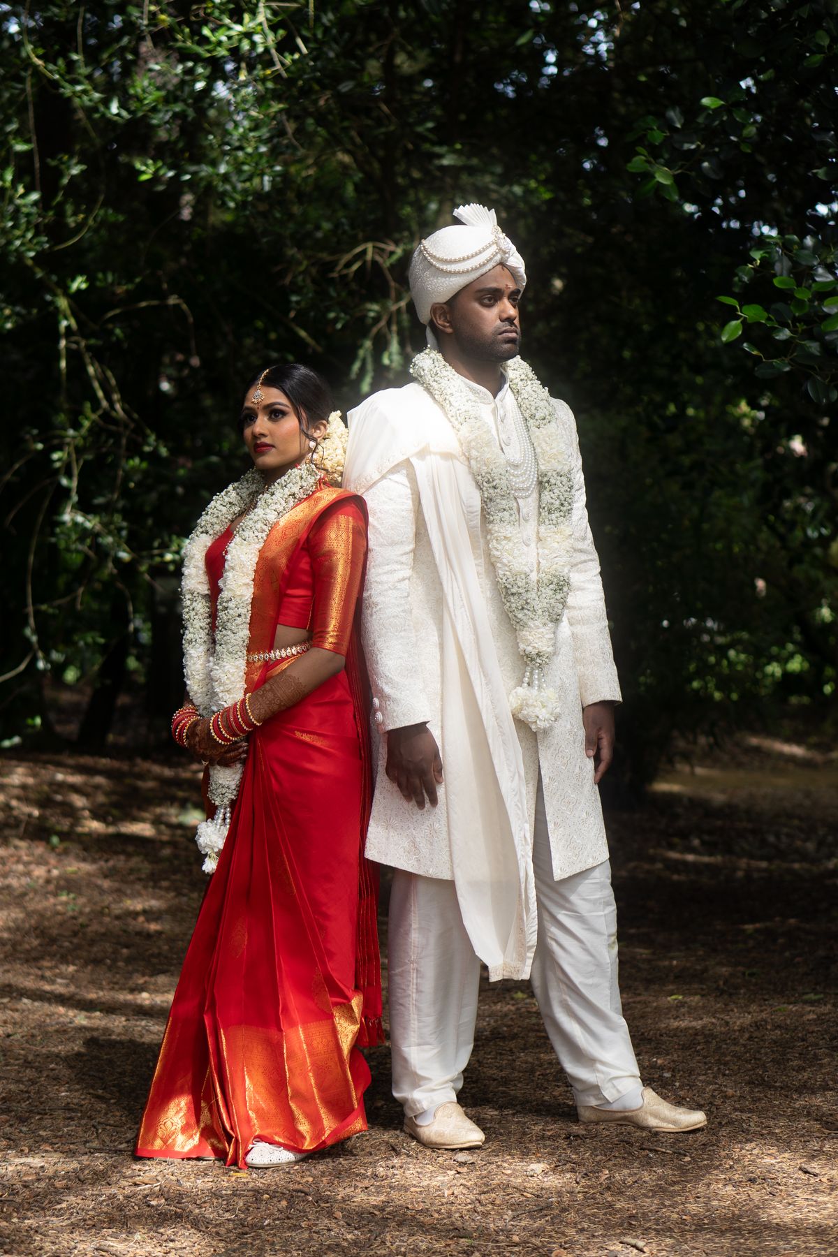 A Hindu bride in a red silk saree and a groom in an ivory sherwani and turban stand back to back, both wearing white flower garlands, in dappled woodland light