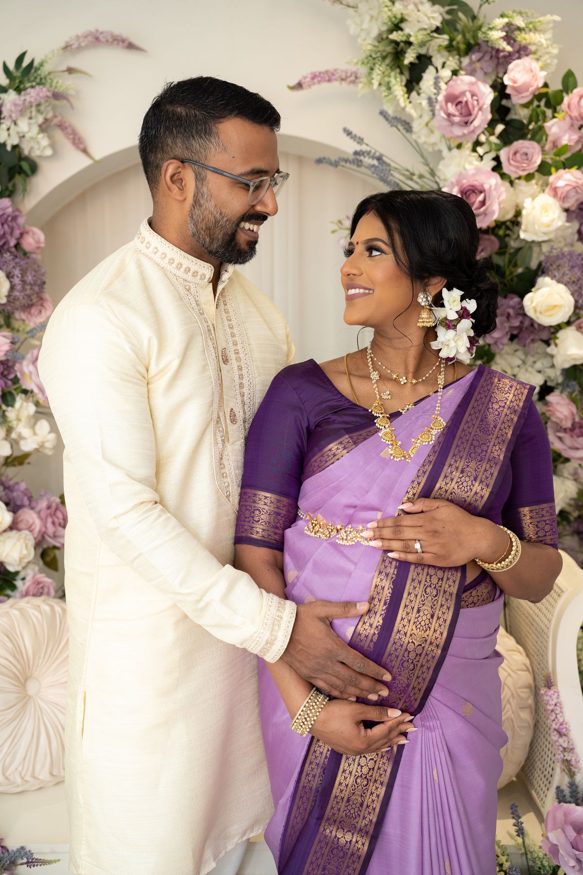 An expectant couple smile at each other in front of a floral arch, her hands resting on her bump in a lilac and purple silk saree